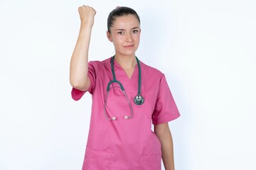 Fierce young caucasian doctor woman wearing pink uniform over white background holding fist in front as if is ready for fight or challenge, screaming and having aggressive expression on face.