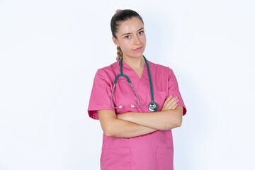 Waist up shot of  self confident young caucasian doctor woman wearing pink uniform over white background has broad smile, crosses arms, happy to meet with colleagues.