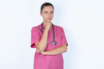 young caucasian doctor woman wearing pink uniform over white background with hand under chin and looking sideways with doubtful and skeptical expression, suspect and doubt.