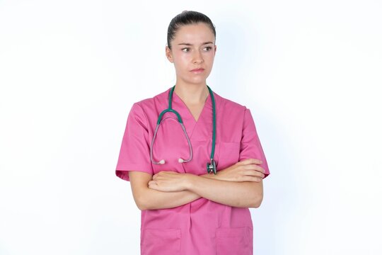 Image of upset young caucasian doctor woman wearing pink uniform over white background with arms crossed. Looking with disappointed expression aside after listening to bad news.