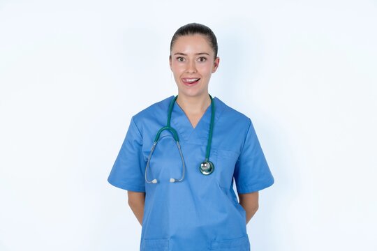 Funny young caucasian doctor woman wearing medical uniform over white background makes grimace and crosses eyes plays fool has fun alone sticks out tongue.