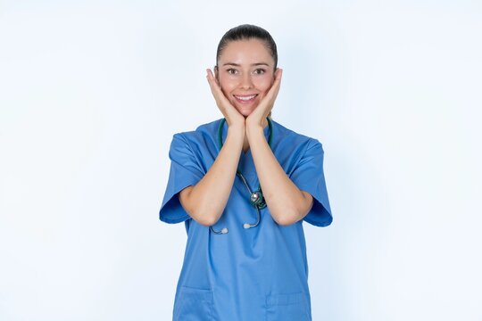 Happy young caucasian doctor woman wearing medical uniform over white background touches both cheeks gently, has tender smile, shows white teeth, gazes positively straightly at camera,