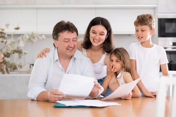 Fototapeta premium Positive husband is sitting at kitchen and reading document about career advancement at work, moneymaking financial report. Smiling wife and happy children hug successful dad