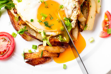 Fried potatoes with egg with liquid flowing yolk, herbs and tomatoes on a white plate.