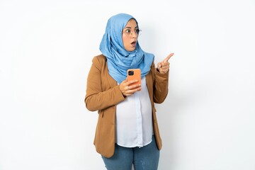 Stunned Young beautiful pregnant muslim woman wearing hijab over white background points sideways...