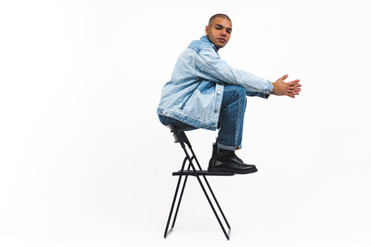 Young black guy in denim clothing sits on top of chair against white background. High quality photo