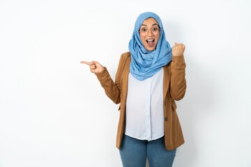 Young beautiful pregnant muslim woman wearing hijab over white background points at empty space holding fist up, winner gesture.