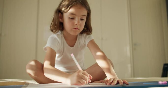 A girl writes letters in a notebook on the floor at home. The child studies and learns lessons. A Caucasian child is studying alone in the living room. High quality 4k footage