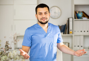 Fototapeta premium Portrait of positive man physician wearing surgical scrubs standing in his office and explaining with gestures.