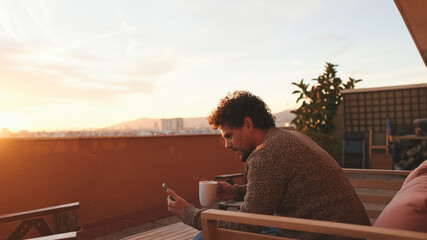 Happy guy sitting on the terrace drinks coffee, browses the web on smartphone