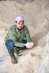 Experienced positive young bearded farmer holding handful of soy flour and checking quality while squatting near pile of feed in livestock farm storage