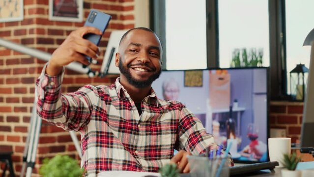 Joyful cheerful african american man taking selfie while smiling and showing thumbs up sign, buttoning up shirt. Person in modern house capturing photo using smartphone with background tv noise