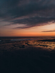 Sunset at the beach. The sky is orange and blue, with pink and purple clouds. Waves crash on the shoreline, reflecting the sun on the wet sand.