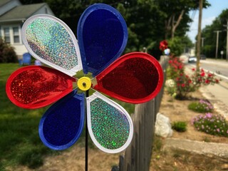 Colorful Spring and Summer Patriotic American Red, White, and Blue Plastic Pinwheel Spinner Toy in foreground House and Road in Background Windmill waiting for Wind Symbol of freedom and childhood.