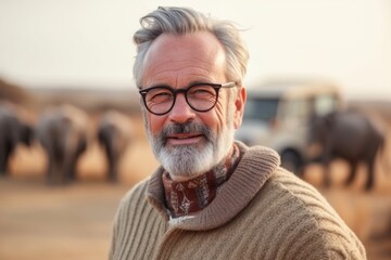 Portrait of senior man with grey hair and eyeglasses standing in front of herd of elephants