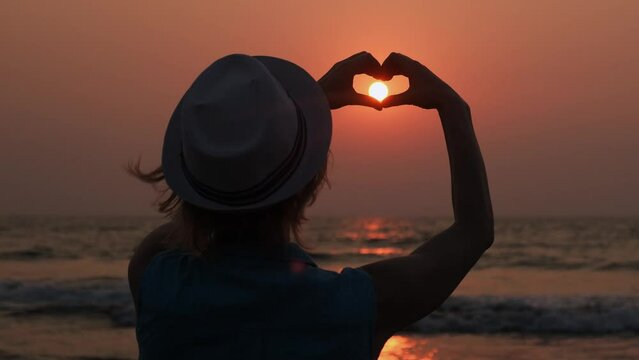 Back view of a young woman wearing hat making heart shaped hands against the beautiful sunset on the sea. Female silhouette on the setting sun background.