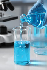 Scientist pouring light blue liquid from flask into beaker in laboratory, closeup