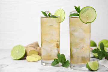 Glasses of tasty ginger ale with ice cubes and ingredients on white marble table