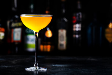 Golden alcoholic cocktail drink with whiskey, apple liqueur, ginger syrup, lemon juice, green basil and ice, dark bar counter background