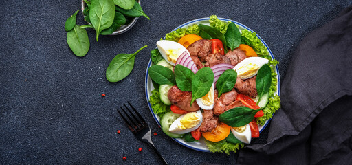 Perigord salad with roasted chicken liver, red tomatoes, cucumbers, boiled eggs, lettuce and spinach. Black kitchen table background, top view