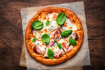 Pizza with spicy salami sausage, mozzarella cheese, tomato sauce and green basil on rustic wooden kitchen table background, top view