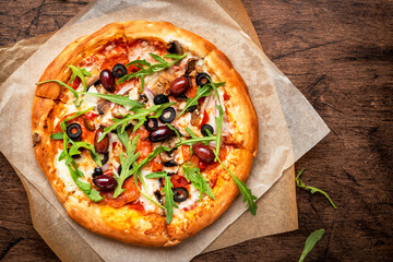 Pizza with salami sausage, mozzarella cheese, mushrooms, black olives, spicy tomato sauce and arugula, rustic wooden table background, top view