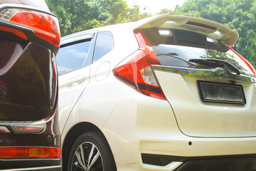 low angle view of back side of white honda jazz car parked in a row in outdoor area and beside of red car bumper with green trees