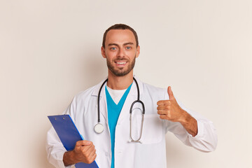 Friendly young male doctor with beard stands on white background wearing white coat with stethoscope on his neck and clipboard in hand, thumb up and smiles, professional people concept, copy space