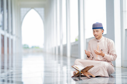  An Asian Muslim man is sitting and reading the Quran. The peace in the mosque makes it an energetic atmosphere of faith, with copy space,  Arabic word translation: The Holy Al Quran.