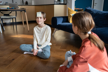 Smiling teen sister and child brother play together who am i guess sticker game sit on floor at...