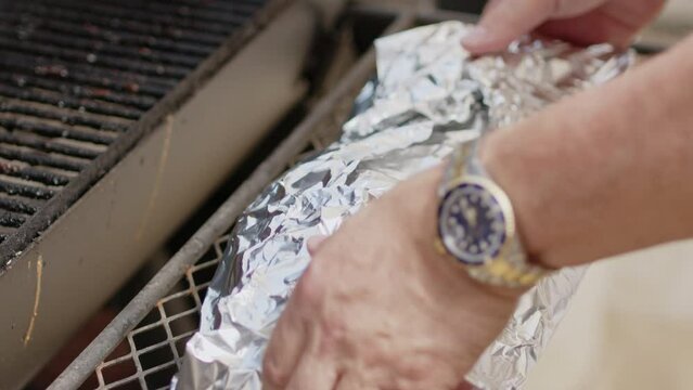 Man foil wrapping BBQ pork ribs and placing them on hit pit smoker with chicken thighs. Slow motion