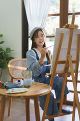 Young female painter holds color palette and use brush to painting artwork on canvas in home studio