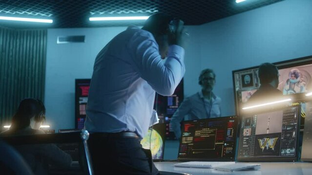 African American flight control employee puts on headset, sits down at computers, monitors space mission in mission control center. Team watch space rocket launch on big digital screens on background.