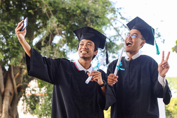 Happy Asian males college graduates in caps and gowns taking selfie on graduation day
