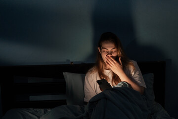 Tired brunette woman yawning and covering her mouth while lying in bed and browsing social media on smart phone at night at home