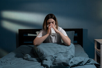 Sick young woman in bed blowing nose in napkin at night. Lady suffering cold and flu having...