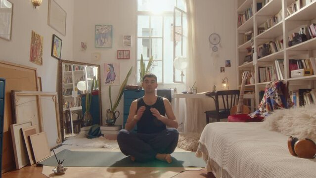 Young barefoot man wearing loose pants walking in sunlit room, sitting in lotus pose on mat, doing Namaste greeting, then meditating with hands in mudras and eyes closed. Zoom shot