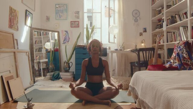 Young black woman sitting with eyes closed in lotus pose on mat, holding hands in mudras, practicing yoga meditation at home in cozy room with aroma incense sticks. Zoom shot