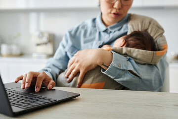 Mom holding her baby and working online on laptop at table in the kitchen - Powered by Adobe