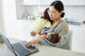 Asian young mother with two kids working online on laptop while sitting in the kitchen