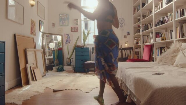 Young beautiful black girl wearing pareo putting on headphones, turning on music on smartphone, then dancing barefoot in sunlit living room. Full length shot