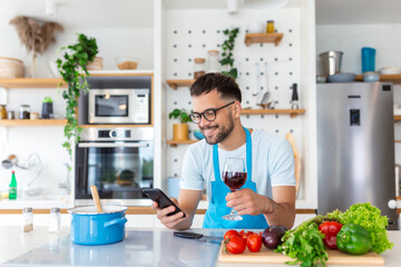 Happy young man preparing romantic dinner searching vegetable recipes diet menu cookbook app using smartphone, smiling husband holding phone cooking healthy vegan food cut salad in kitchen interior