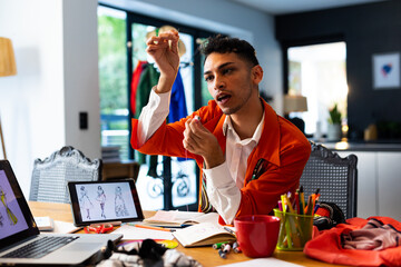 Biracial transgender fashion designer holding thread, using laptop and tablet