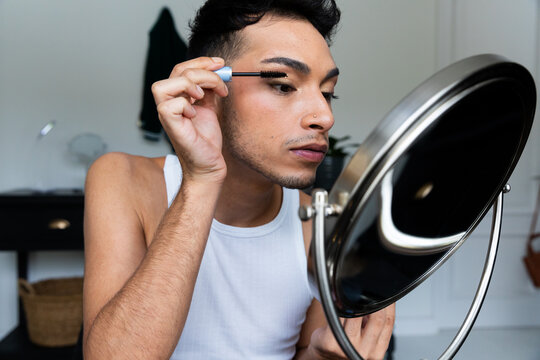 Biracial transgender man looking in mirror and putting on make-up, applying mascara - Powered by Adobe