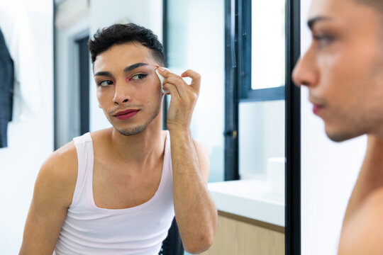 Happy biracial transgender man looking in mirror, using cotton pad for cleansing face in bathroom