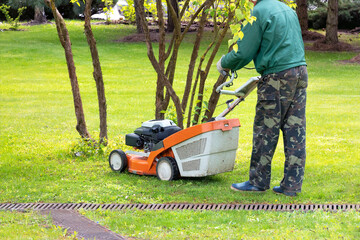 A gardener in camouflage clothing mows a grassy lawn with a petrol lawn mower on a spring day.