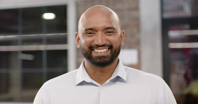 Portrait of happy biracial bald creative businessman with beard smiling in office, in slow motion