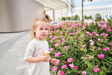 Obraz na płótnie Canvas A hungry child enjoys eating ice cream outdoors. Portrait of a cute baby with a dirty face licking a creamy ice cream while walking in a city park, outdoor activities.