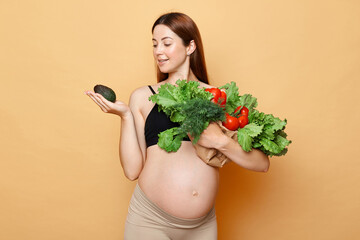 Expecting mother holding avocado. Healthy eating during pregnancy. Pregnant woman embraces fresh vegetables posing isolated over beige background