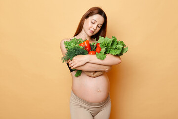 Charming brown haired pregnant woman embraces fresh vegetables posing isolated over beige background enjoying her pregnancy taking care of her and baby health.